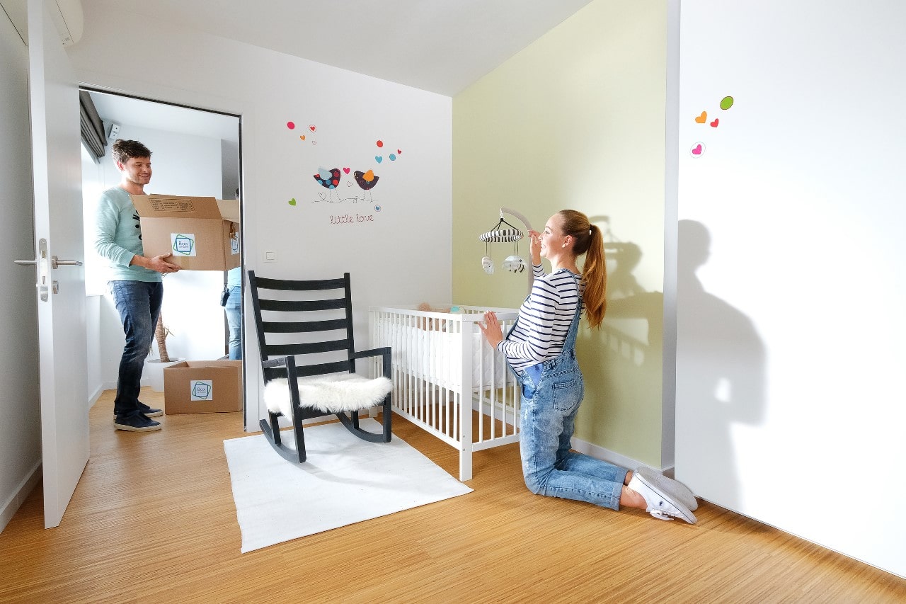 Jongeman staat in de kinderkamer en draagt een verhuisdoos van Box@Home terwijl zijn vriendin het babybed aankleedt