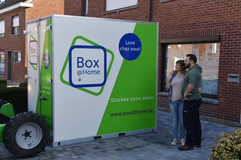 A young couple stand in front of their house and look with satisfaction at their newly delivered Box@Home storage space